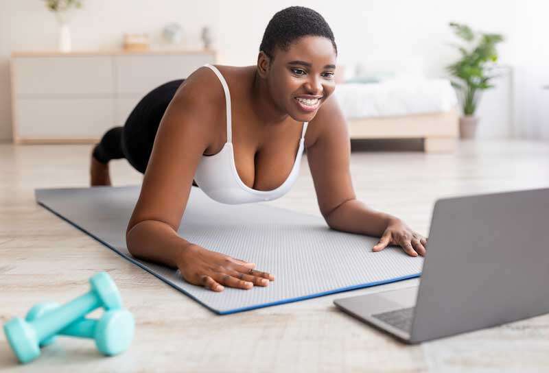 black lady exercising at home, following personal trainer's online instructions on laptop, standing in plank pose